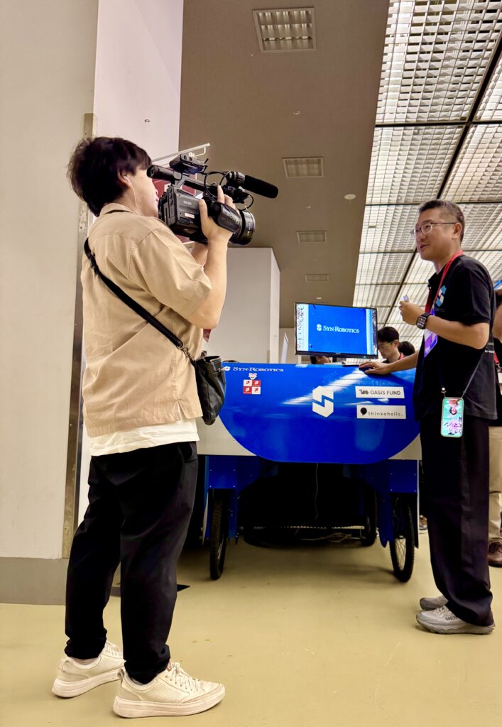 IVS会場のシンロボブース前でカメラを構える取材スタッフと取材に応じるシンロボ代表の柳本